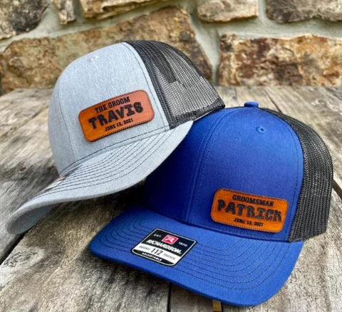 Two personalized leather-patch trucker hats for groomsmen, one reading The Groom and one Groomsman, resting on a rustic wood table.
