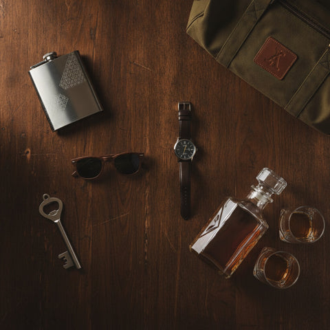 A flat-lay of personalized groomsmen gifts across budgets on dark walnut wood: an engraved flask, wood sunglasses, a bottle opener, a personalized watch, a monogrammed canvas duffle, and an engraved whiskey decanter with rocks glasses.