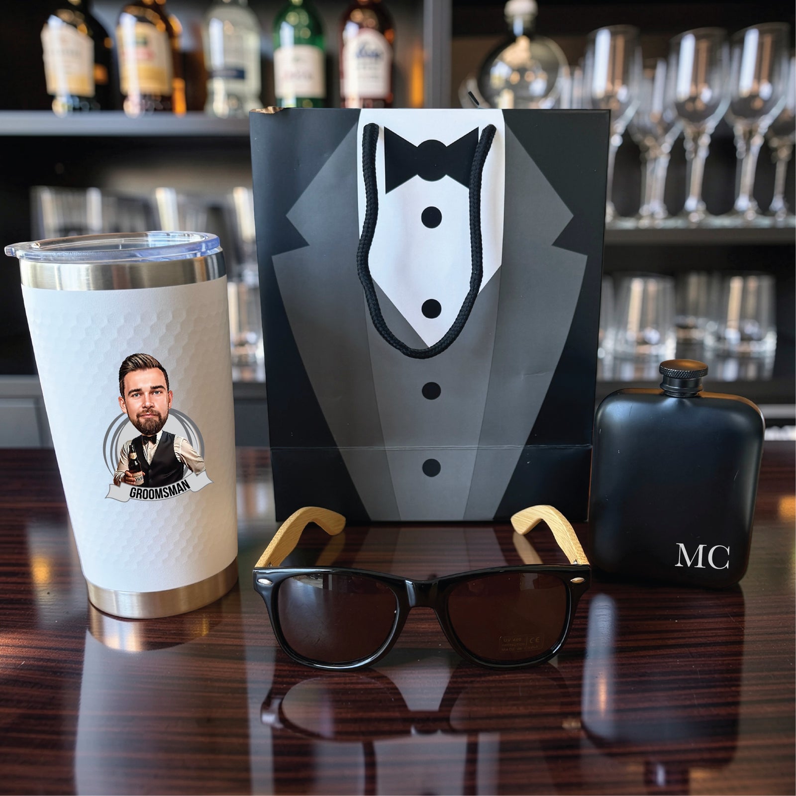 Groomsmen gift set with a caricature tumbler, black flask engraved MC and bamboo sunglasses in front of a tuxedo gift bag on a bar