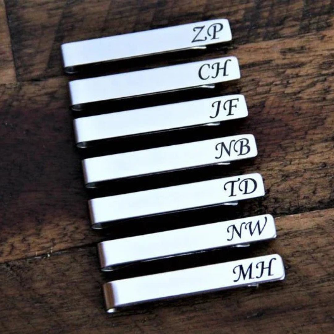 Stack of seven white rectangular cufflinks with black trim, each engraved with sample monogram initials including ZP, CH, JF, NB, TD, NW, and MH, displayed on dark wood surface.