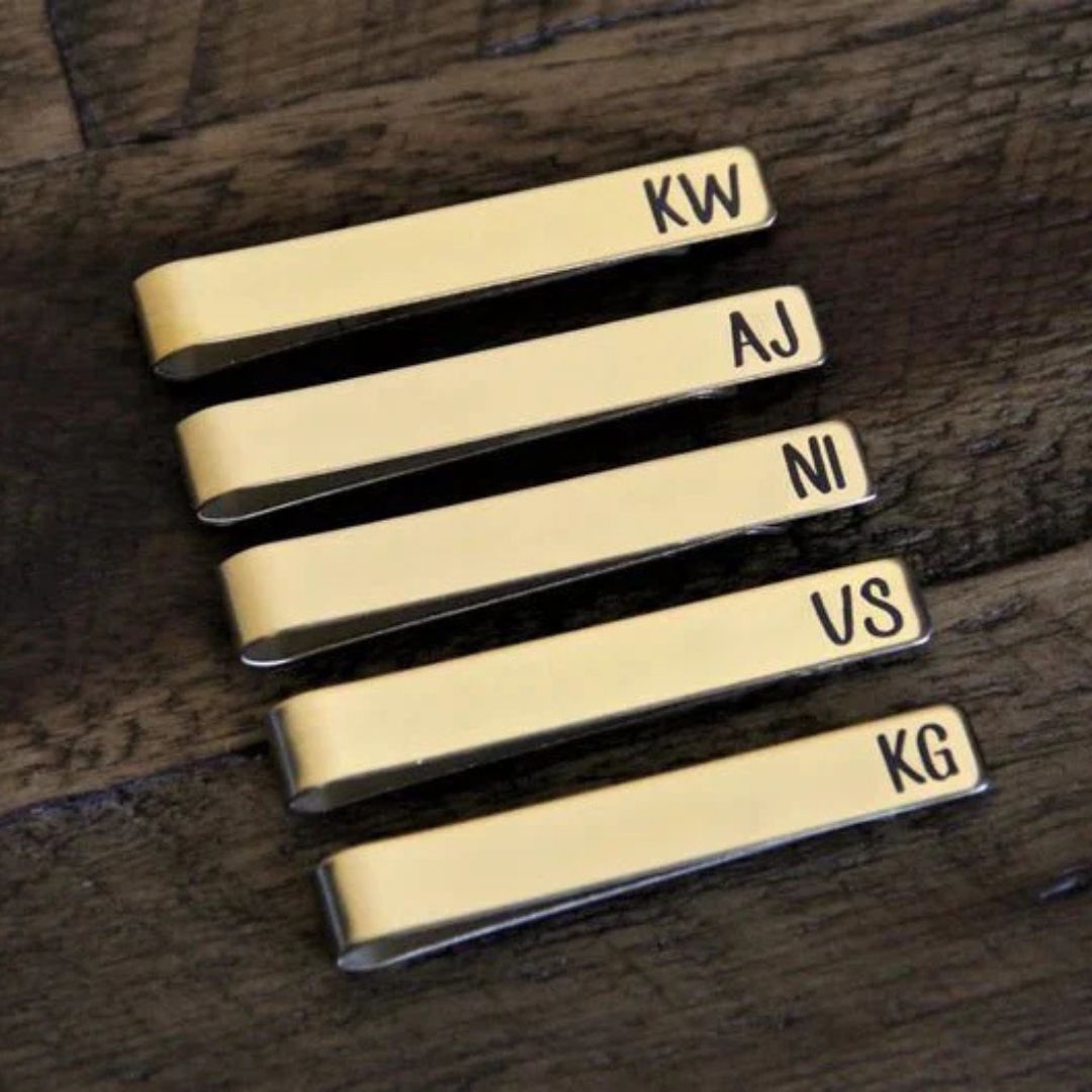 Five gold-tone tie bars stacked vertically, each engraved with sample initials KW, AJ, NI, US, and KG in black lettering
