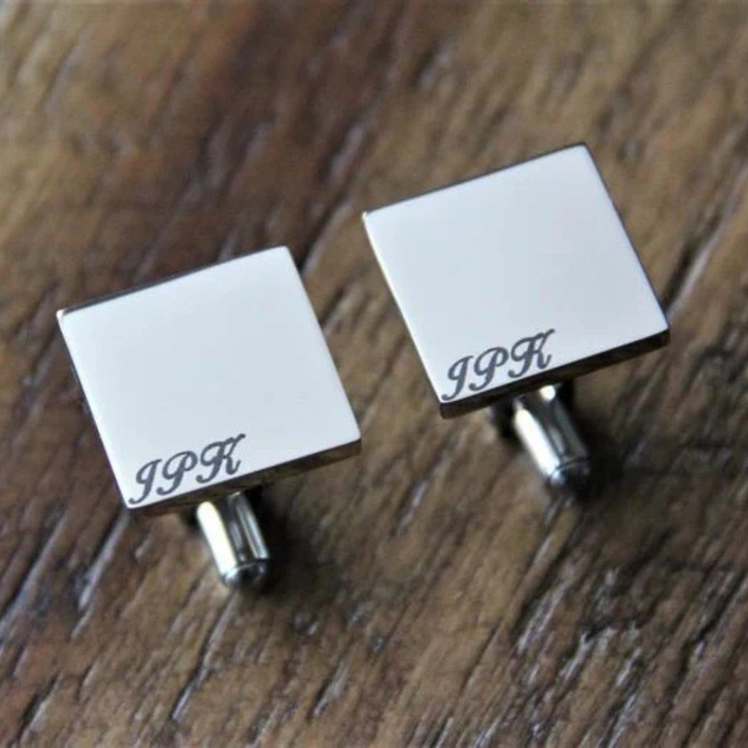 Pair of square silver cufflinks with monogrammed sample initials "JPK" engraved on white enamel face, displayed on wood surface