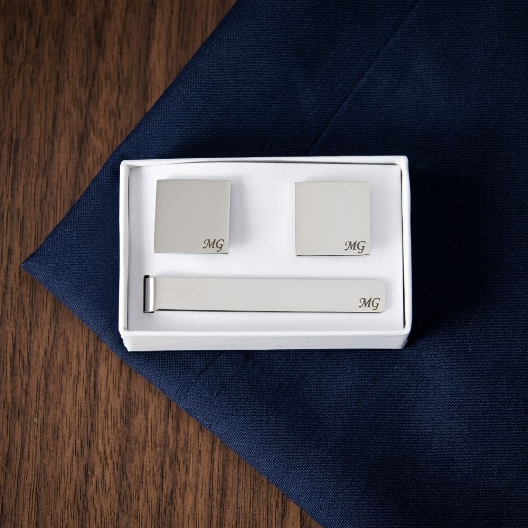 Three-piece cufflinks and tie bar set with monogrammed sample initials "MG" displayed in white gift box packaging on navy fabric background.