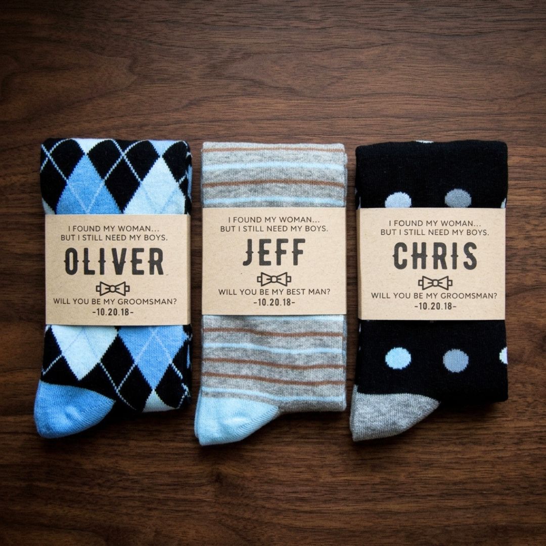 Three personalized socks with kraft paper labels showing sample names Oliver, Jeff, and Chris with groomsman proposal text and bowtie graphic