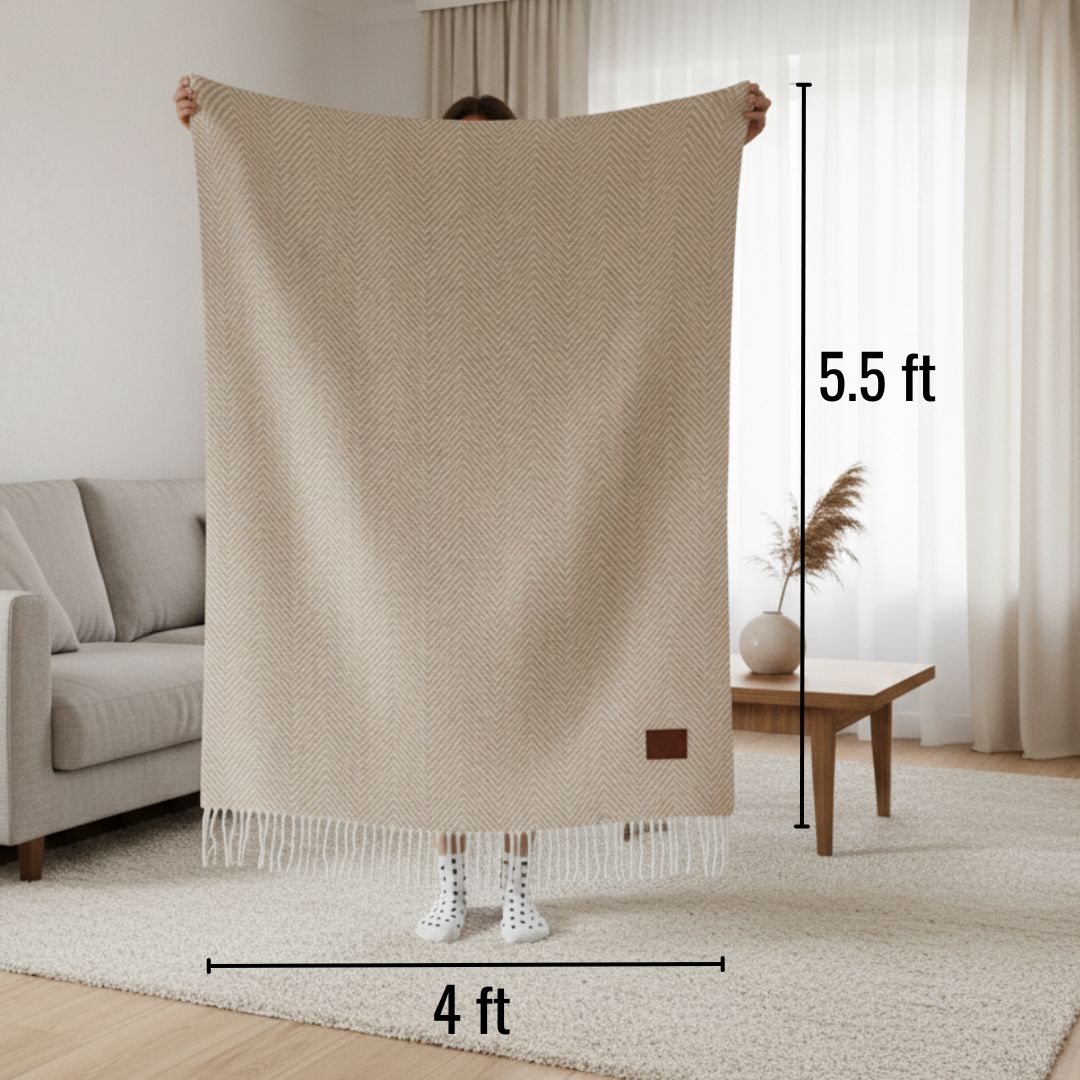 Cream herringbone throw blanket with fringe trim, displayed at 4 feet wide by 5.5 feet tall, featuring a brown patch detail on lower corner