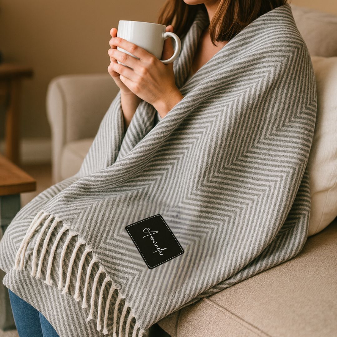 Person wearing grey herringbone throw blanket with fringe trim and black patch featuring sample personalized script text, holding white mug while seated on couch