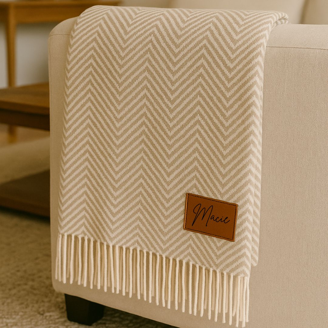 Cream herringbone throw blanket with fringe trim and sample leather patch reading "Marie" in cursive script.