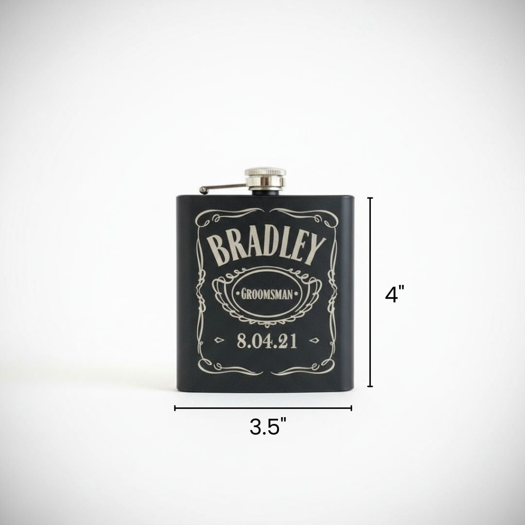 Black stainless steel flask with silver hinged cap, engraved with sample personalization reading "BRADLEY" with "GROOMSMAN" banner and date "8.04.21" in decorative Victorian-style frame