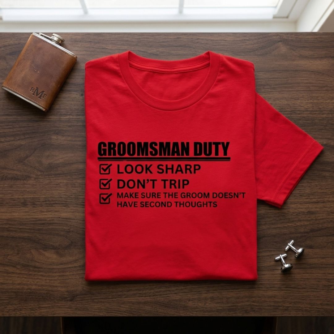 Red t-shirt printed with "Groomsman Duty" checklist including look sharp, don't trip, and make sure groom doesn't have second thoughts, displayed flat with leather flask and cufflinks