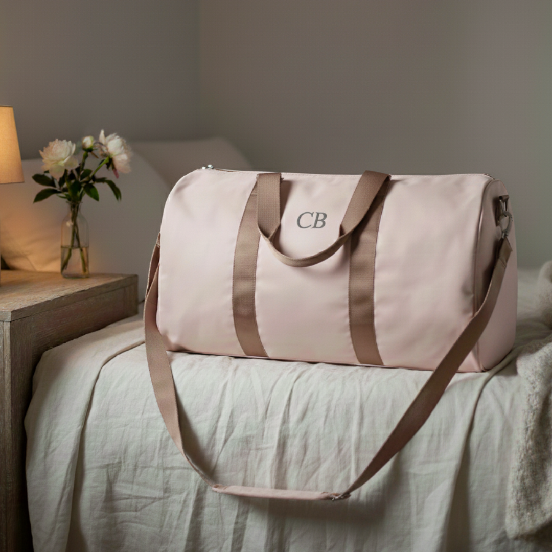 Cream canvas duffle bag with taupe straps, personalized with monogram "CB" on front panel, displayed on bed beside wooden nightstand with flowers