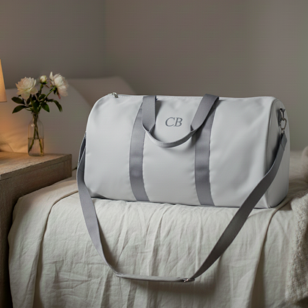 White duffle bag with gray straps and sample monogram "CB" embroidered on front panel, displayed on bed with flowers and bedside table visible