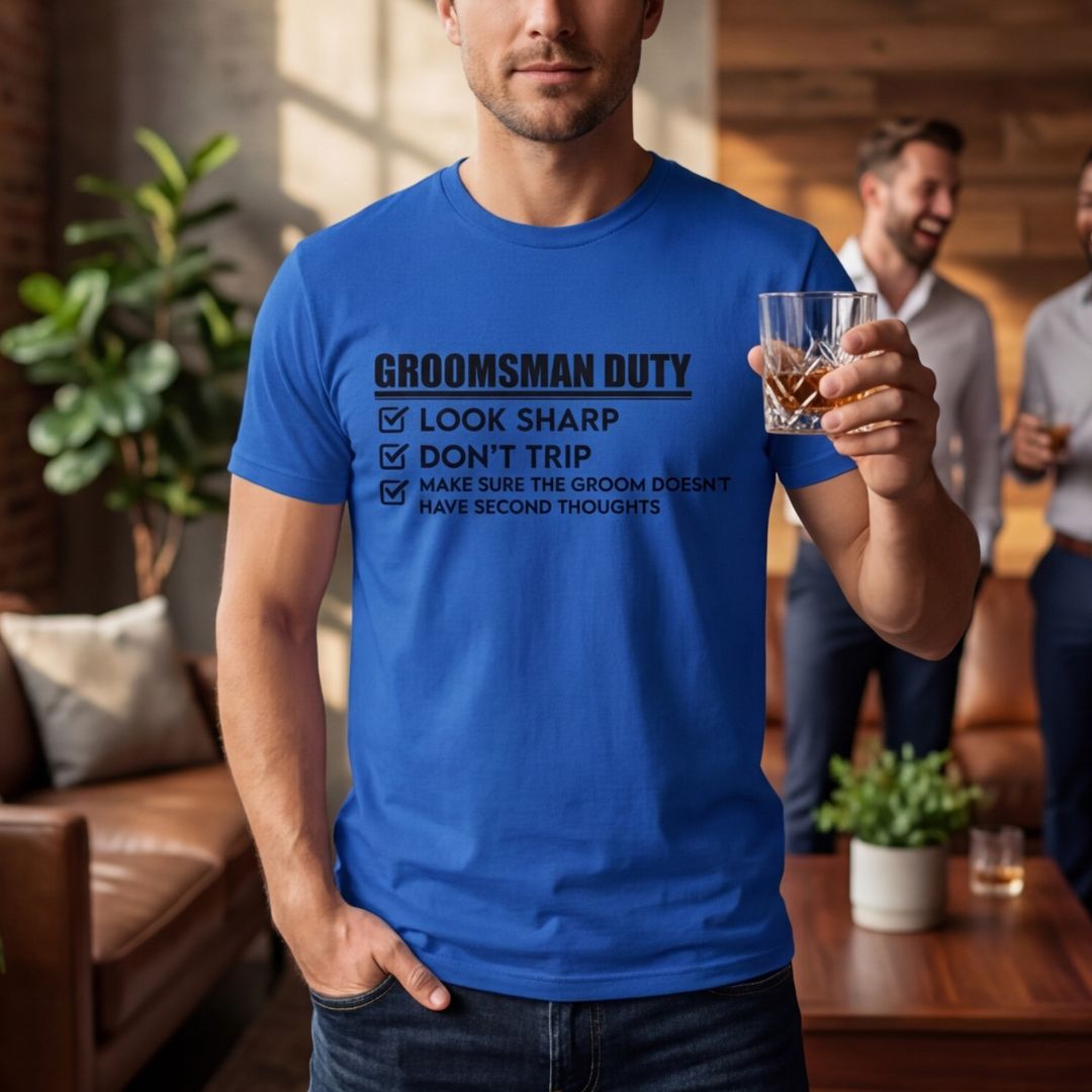 Man wearing royal blue t-shirt printed with "Groomsman Duty" checklist design, holding whiskey glass in lifestyle setting with other guests in background
