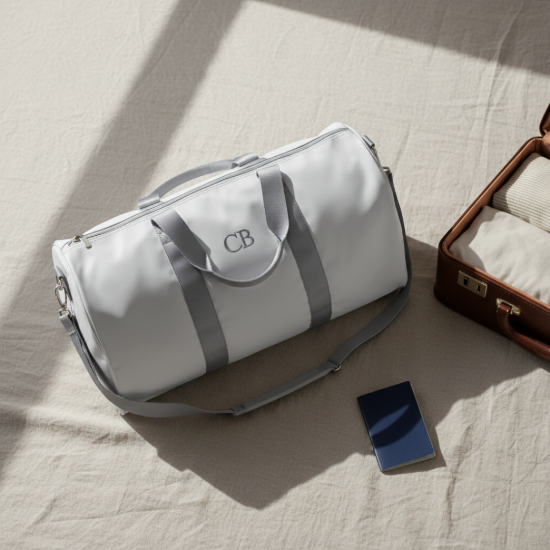 White duffle bag with gray straps and sample monogram "CB" centered on front panel, displayed on light fabric with accessories nearby