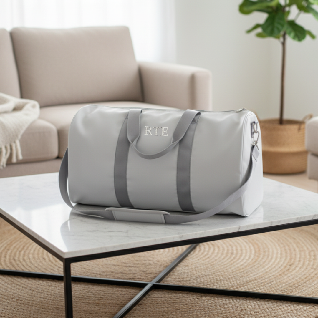 Light gray duffle bag with gray handles and straps, personalized with sample monogram "RTE" in white lettering on front panel, displayed on marble table in modern living room setting.