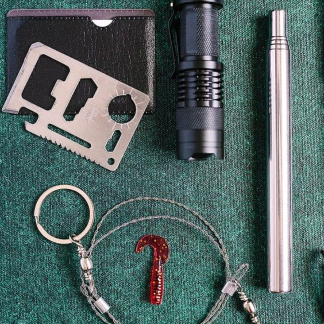 Flatlay arrangement of survival kit contents on green fabric: waterproof case, multi-tool card, black flashlight, silver pen, red carabiner with compass and whistle bracelet, and metal keyring.