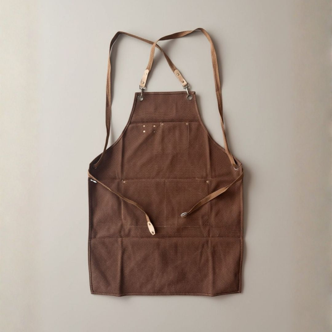 Brown canvas apron with tan leather straps and metal hardware, displayed flat against neutral background with adjustable shoulder straps and multiple front pockets