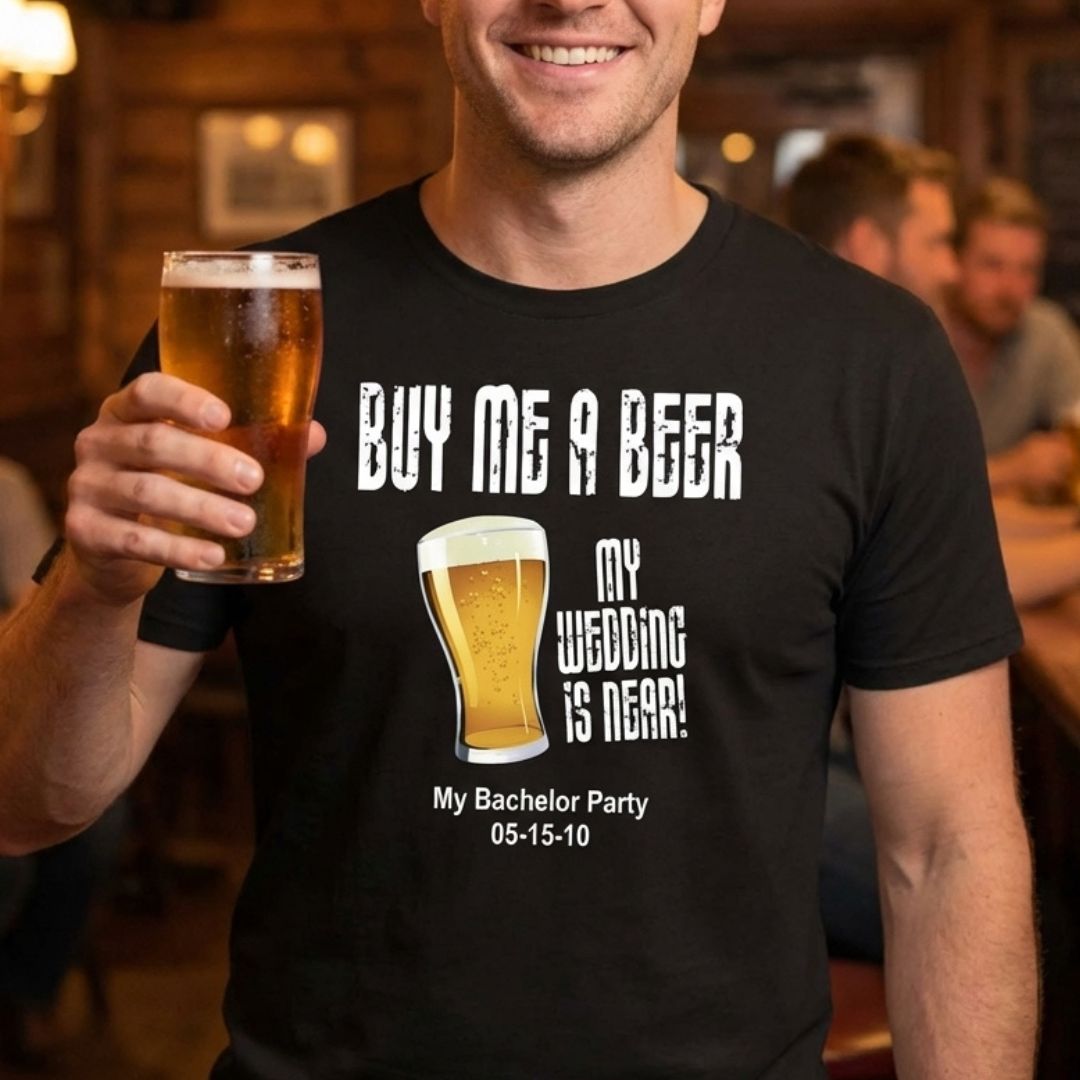 Man wearing black t-shirt printed with "Buy Me a Beer My Wedding Is Near" text, beer glass graphic, and sample date "05-15-10" while holding a glass of beer in a bar setting.