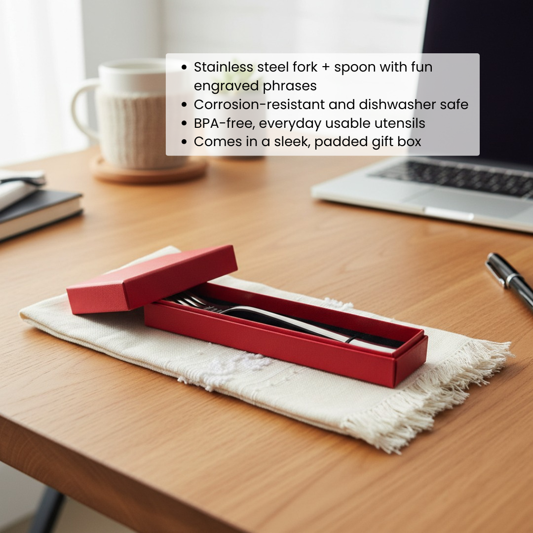 Stainless steel fork and spoon with engraved phrases displayed in open red gift box on cream cloth liner, positioned on wooden desk