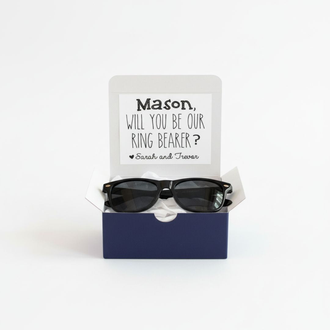 Black wayfarer-style sunglasses nestled in navy gift box with personalized card reading "Mason, will you be our ring bearer?" with sample names Sarah and Trevor