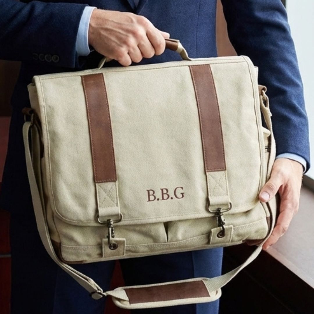 Cream canvas messenger bag with brown leather straps, personalized with sample monogram "B.B.G" on front flap, held by man in navy blazer