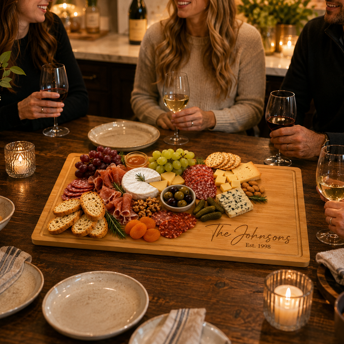 A large wooden charcuterie board engraved with 'The Johnsons Est. 1995' sits on a rustic wooden dining table, surrounded by people holding wine glasses in a warmly lit, cozy kitchen setting.