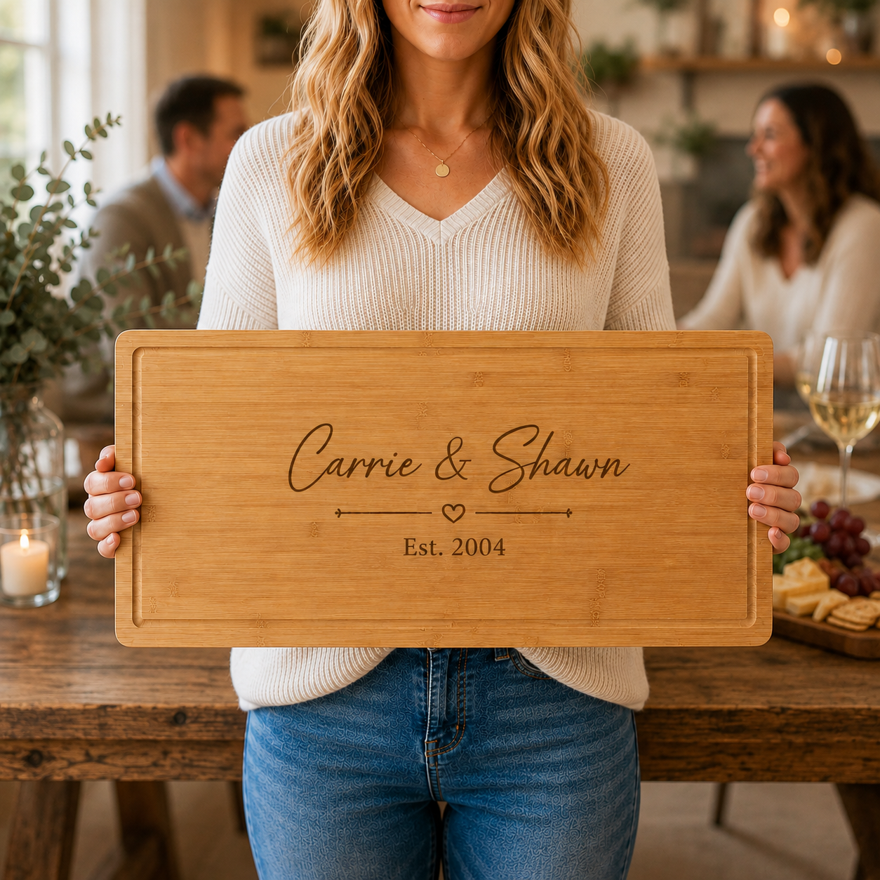Personalized wooden charcuterie board engraved with "Carrie & Shawn" and "Est. 2004" with decorative heart divider, displayed at a gathering with blurred guests in background.