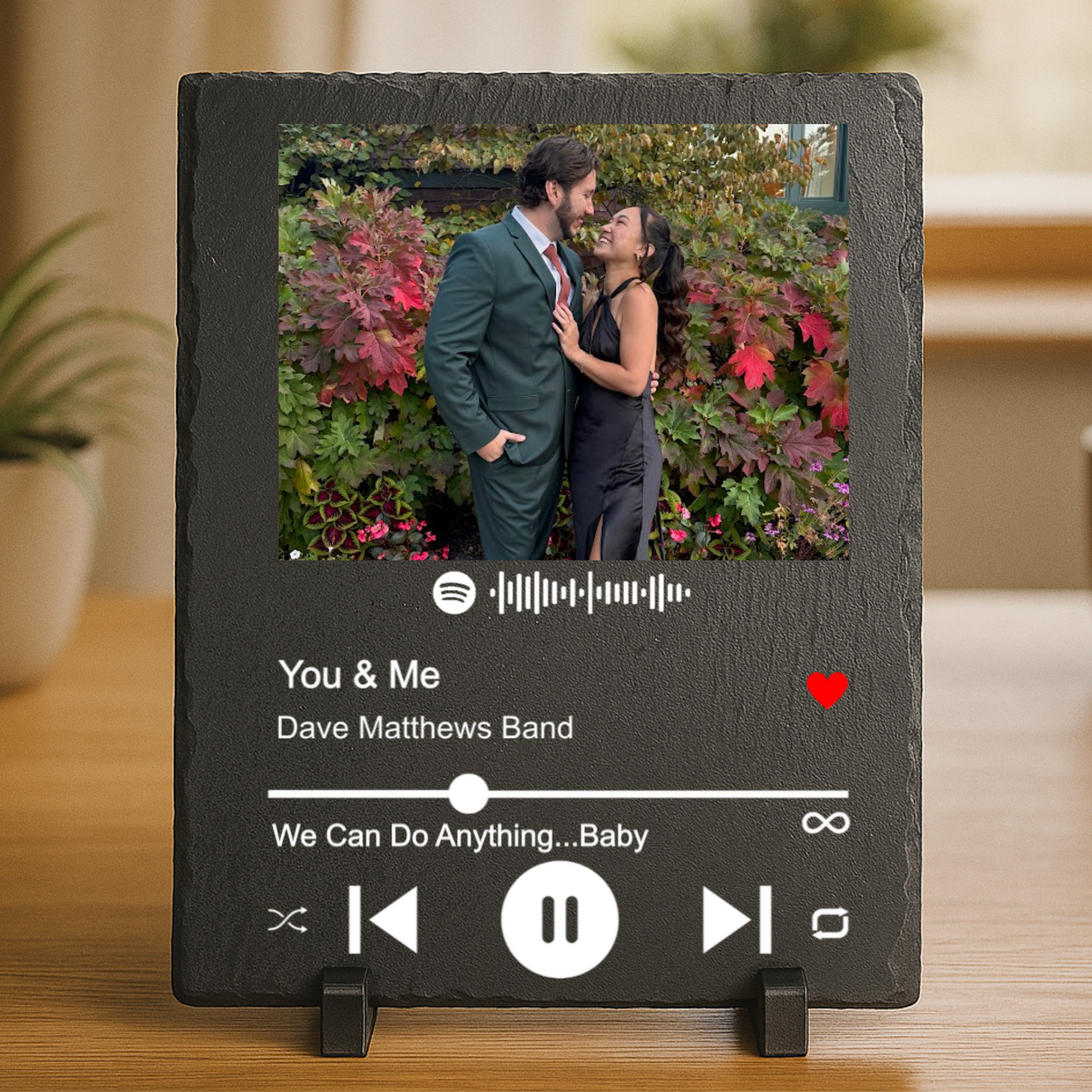 Black slate plaque displaying a customized Spotify player interface with sample photo of couple in front of flowering vines and example song "You & Me" by Dave Matthews Band with playback controls.
