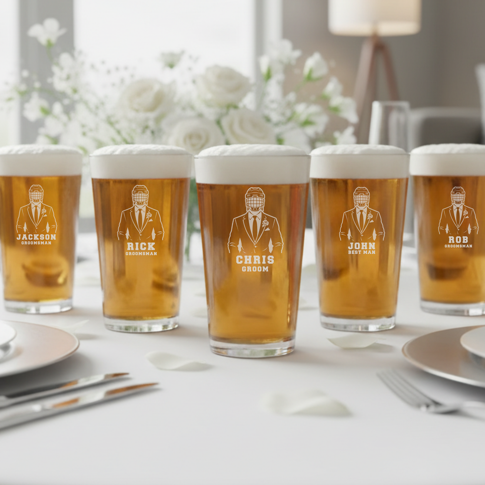 Five personalized pint glasses filled with beer, each laser-engraved with a groomsman figure in hockey helmet and sample names Jackson, Rick, Chris, John, and Rob on a banquet table with white flowers and place settings