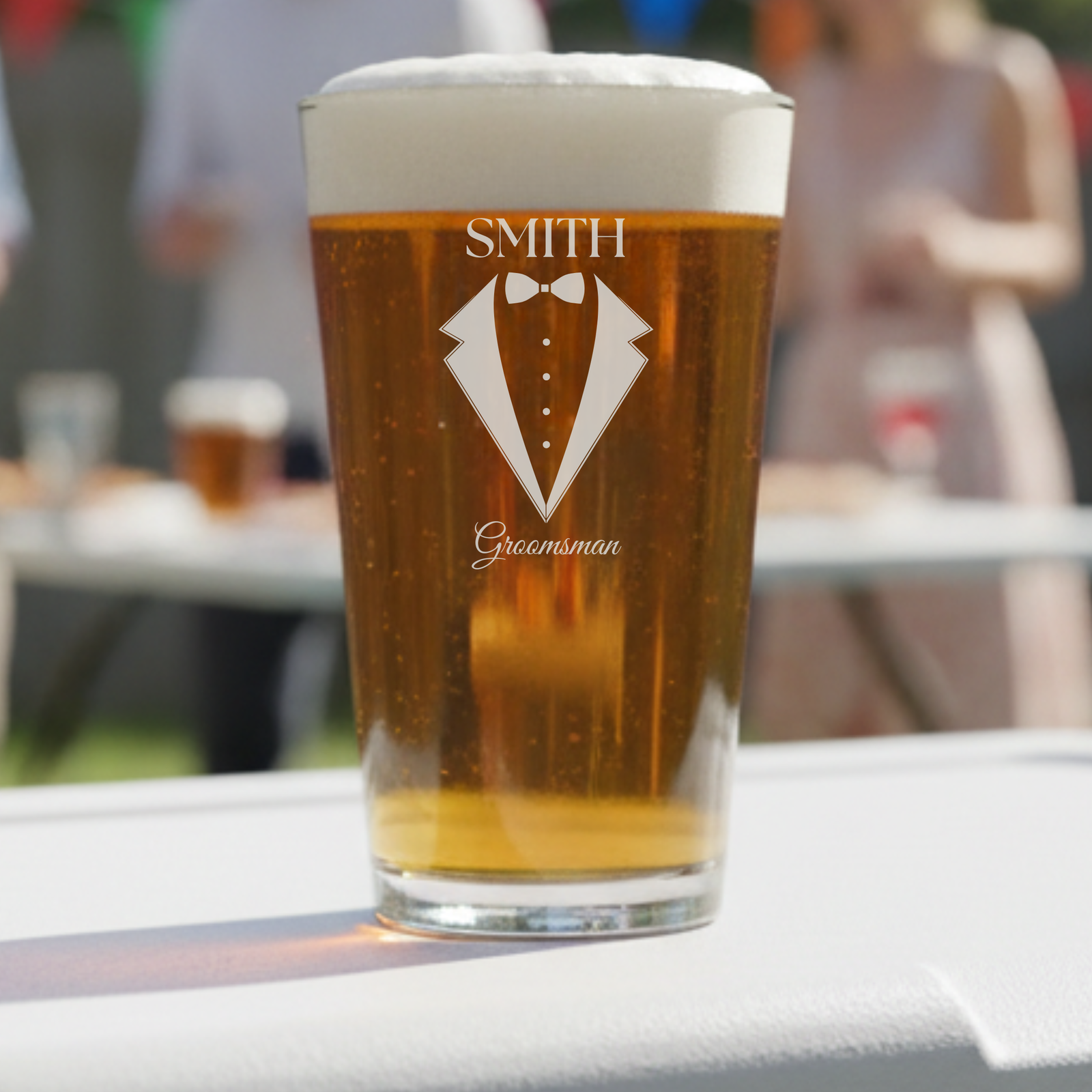 Personalized pint glass filled with beer, engraved with sample name "SMITH" above tuxedo graphic with bowtie and "Groomsman" text below