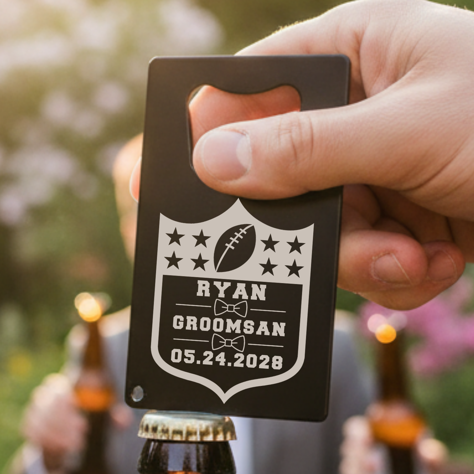Black stainless steel bottle opener with shield design, personalized with sample name "Ryan," football motif, bow-tie icons, and wedding date "05.24.2028," held in hand outdoors.