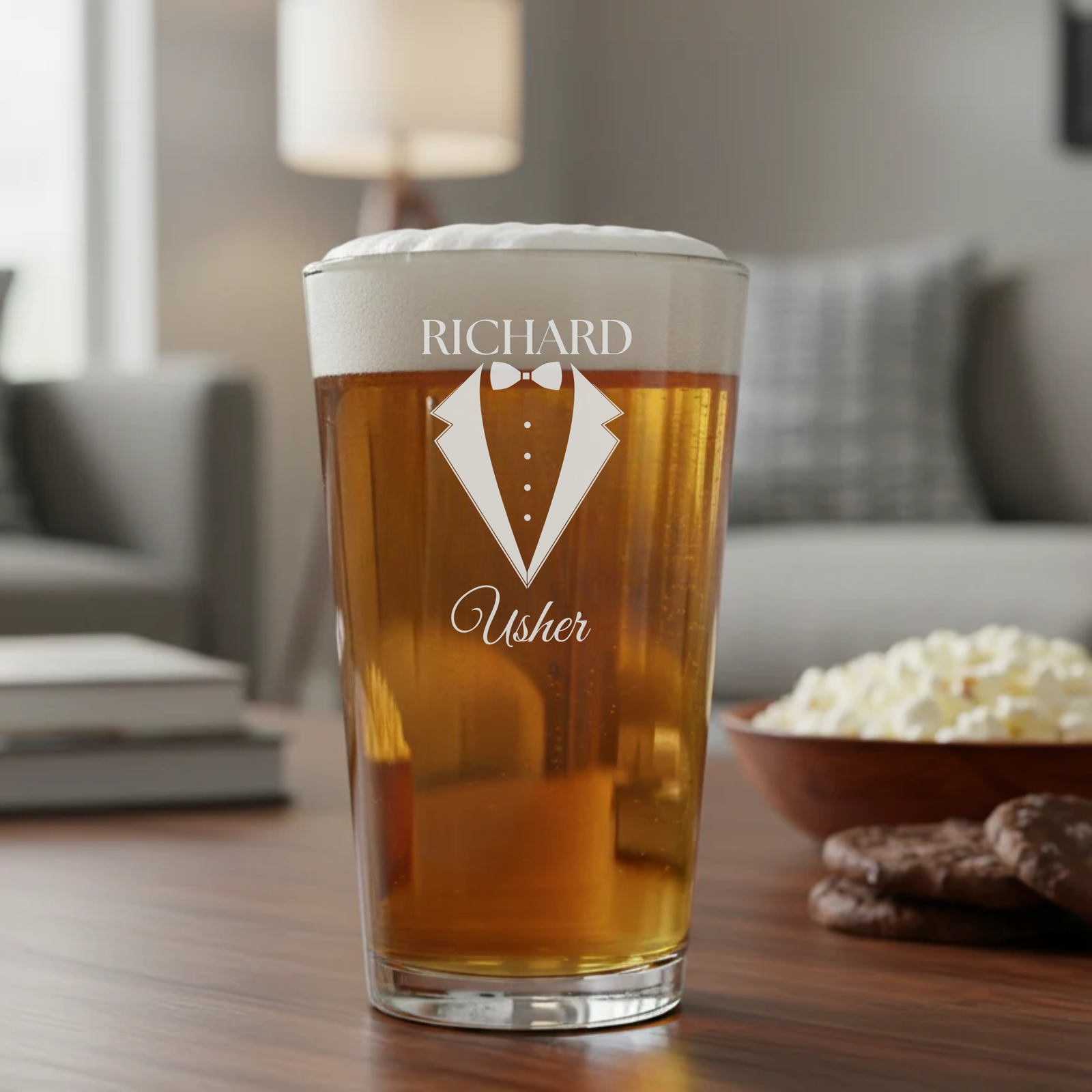Pint glass filled with amber beer, engraved with sample personalization showing "RICHARD" at top, tuxedo with bowtie graphic, and "Usher" text below, displayed on wooden table with home interior background