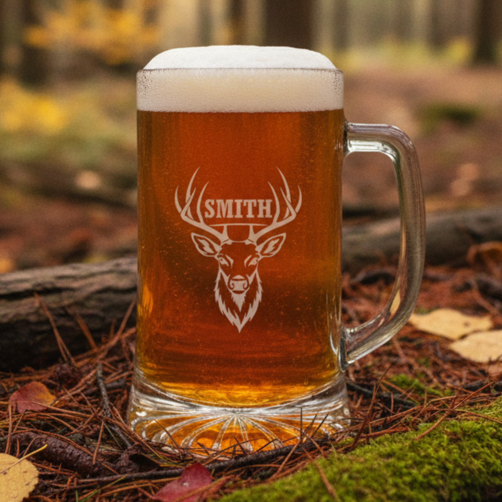 Engraved glass beer mug filled with amber beer and foam, featuring a sample personalization with a deer head design and the name "SMITH" in a forest setting.