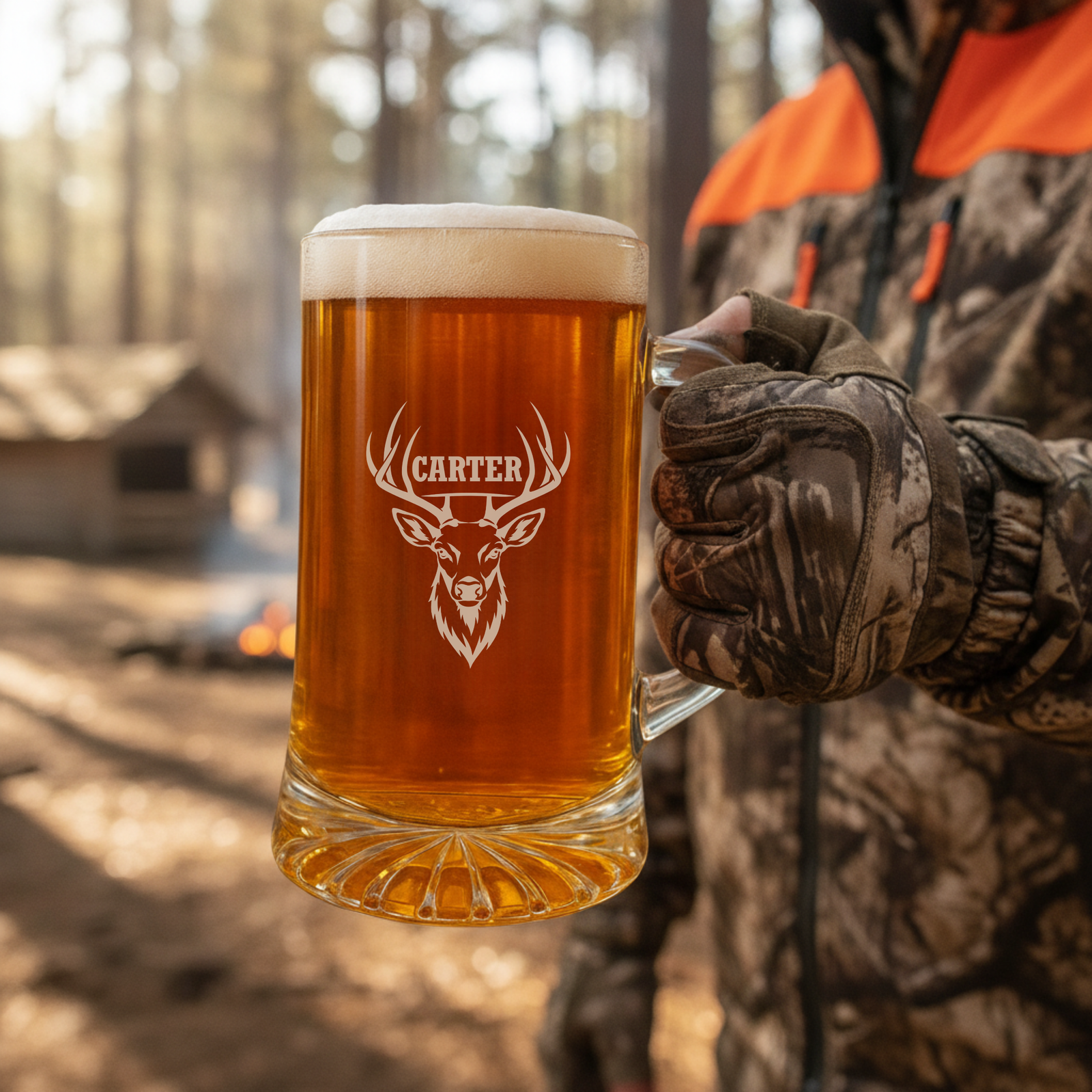 Engraved beer mug with deer head and sample name "Carter" held by person in camouflage outdoors near campfire