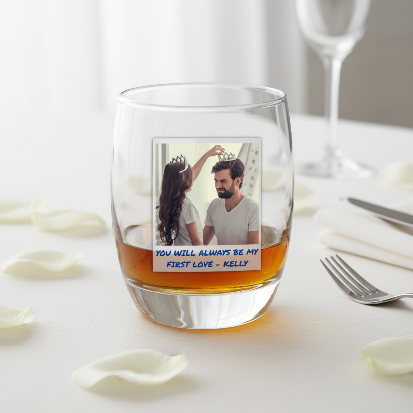 Whiskey glass filled with amber liquid, displaying a custom photo of a couple with crowns and personalized text "YOU WILL ALWAYS BE MY FIRST LOVE - KELLY" printed below the image