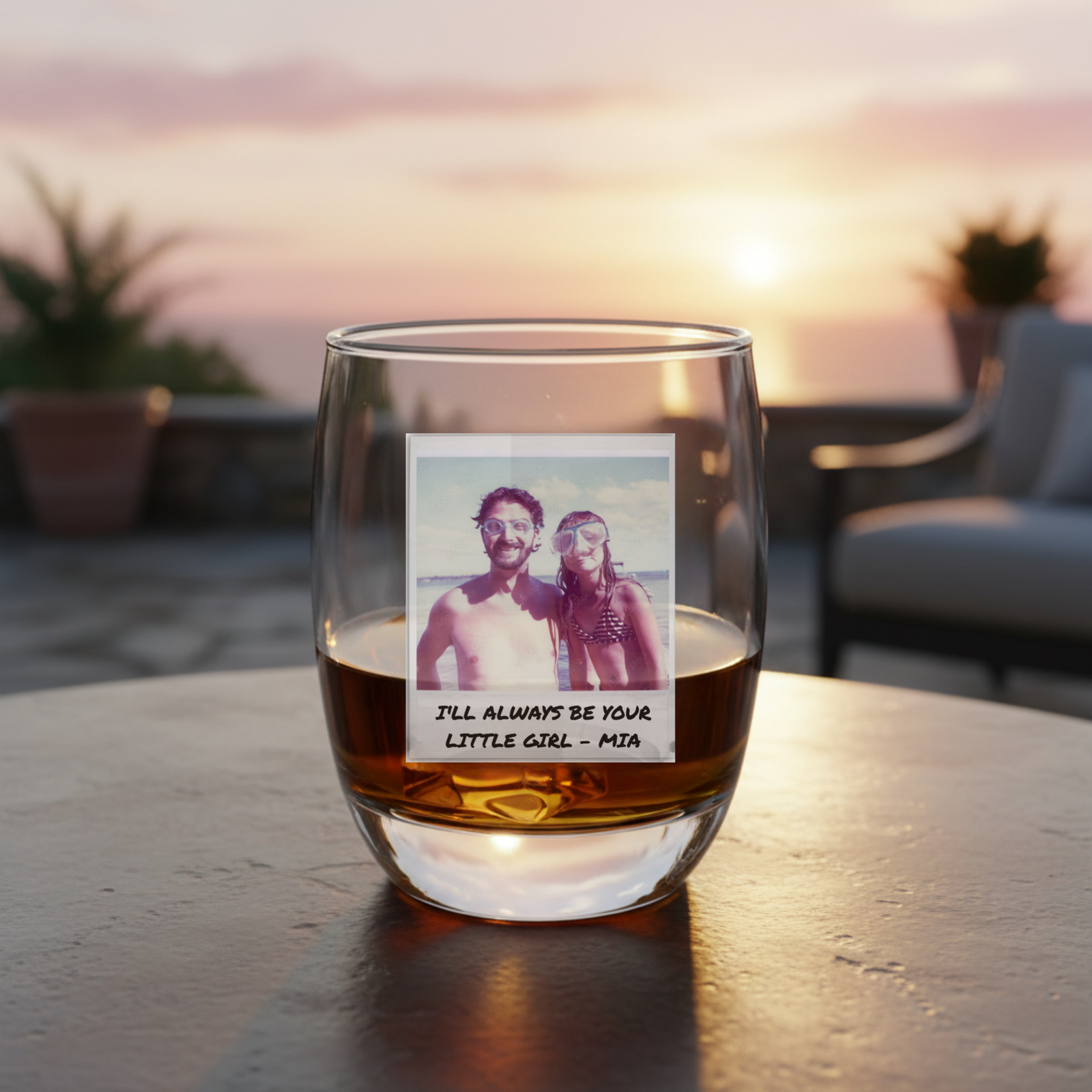 Whiskey glass filled with amber liquid, personalized with sample Polaroid photo of two people and handwritten text reading "I'll Always Be Your Little Girl - Mia", displayed outdoors at sunset