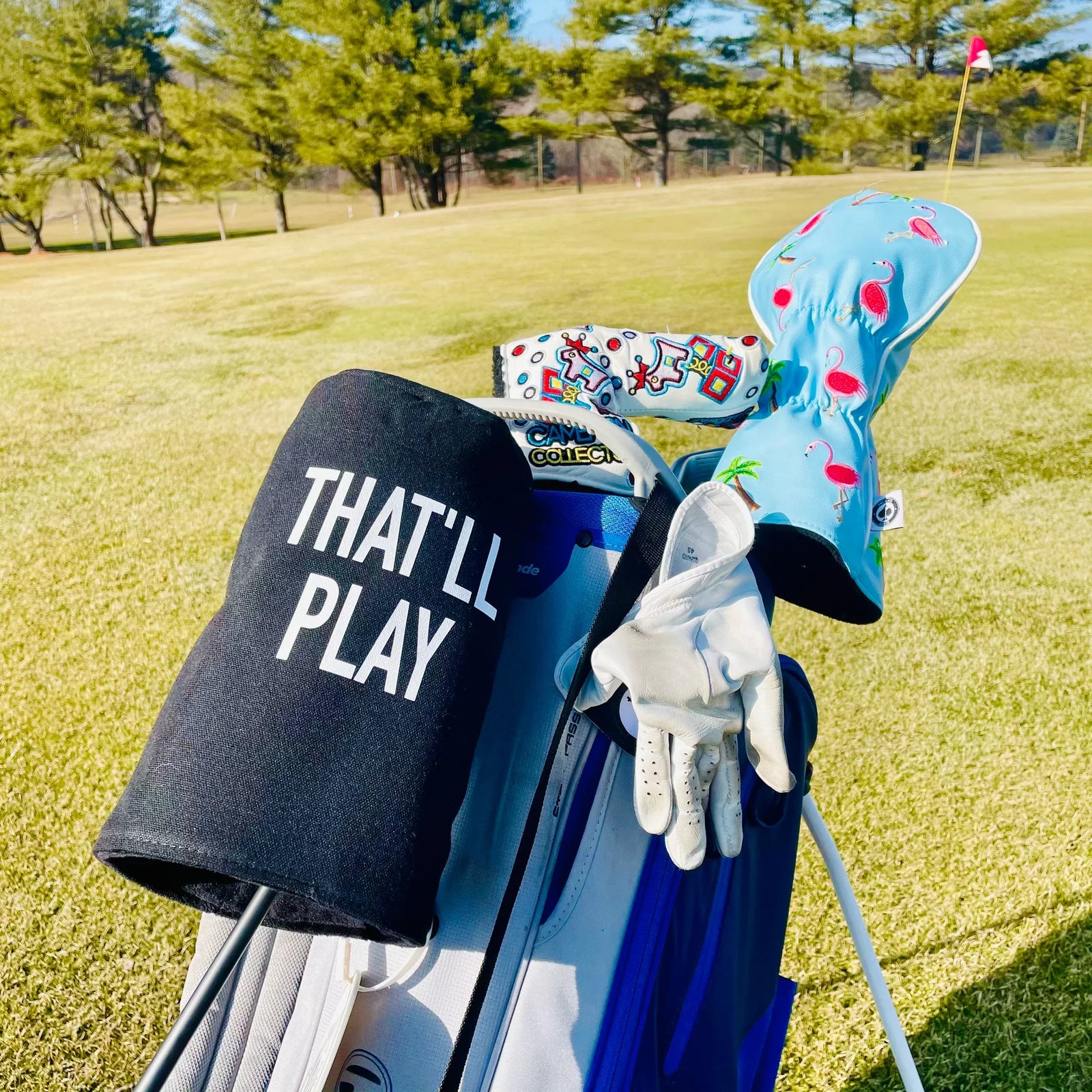 Black golf head cover with "THAT'LL PLAY" text displayed on golf bag alongside colorful patterned head covers on fairway course.