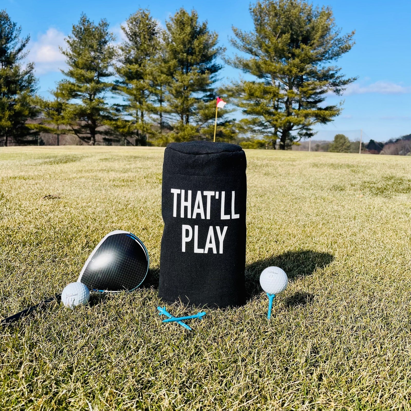 Black barrel head cover printed with "THAT'LL PLAY" text, displayed on golf course with driver, golf balls, and tees
