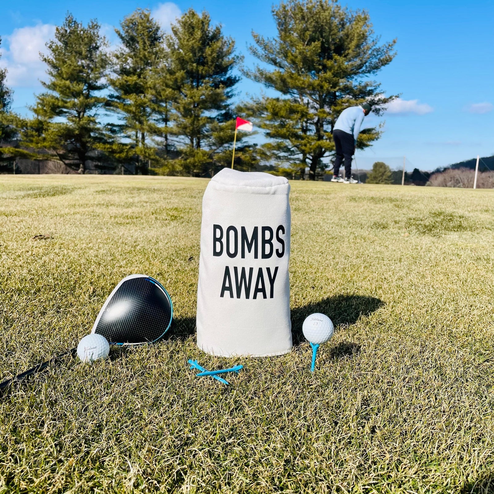 White golf barrel head cover with "BOMBS AWAY" text, displayed on grass next to club, ball, and blue tee on golf course