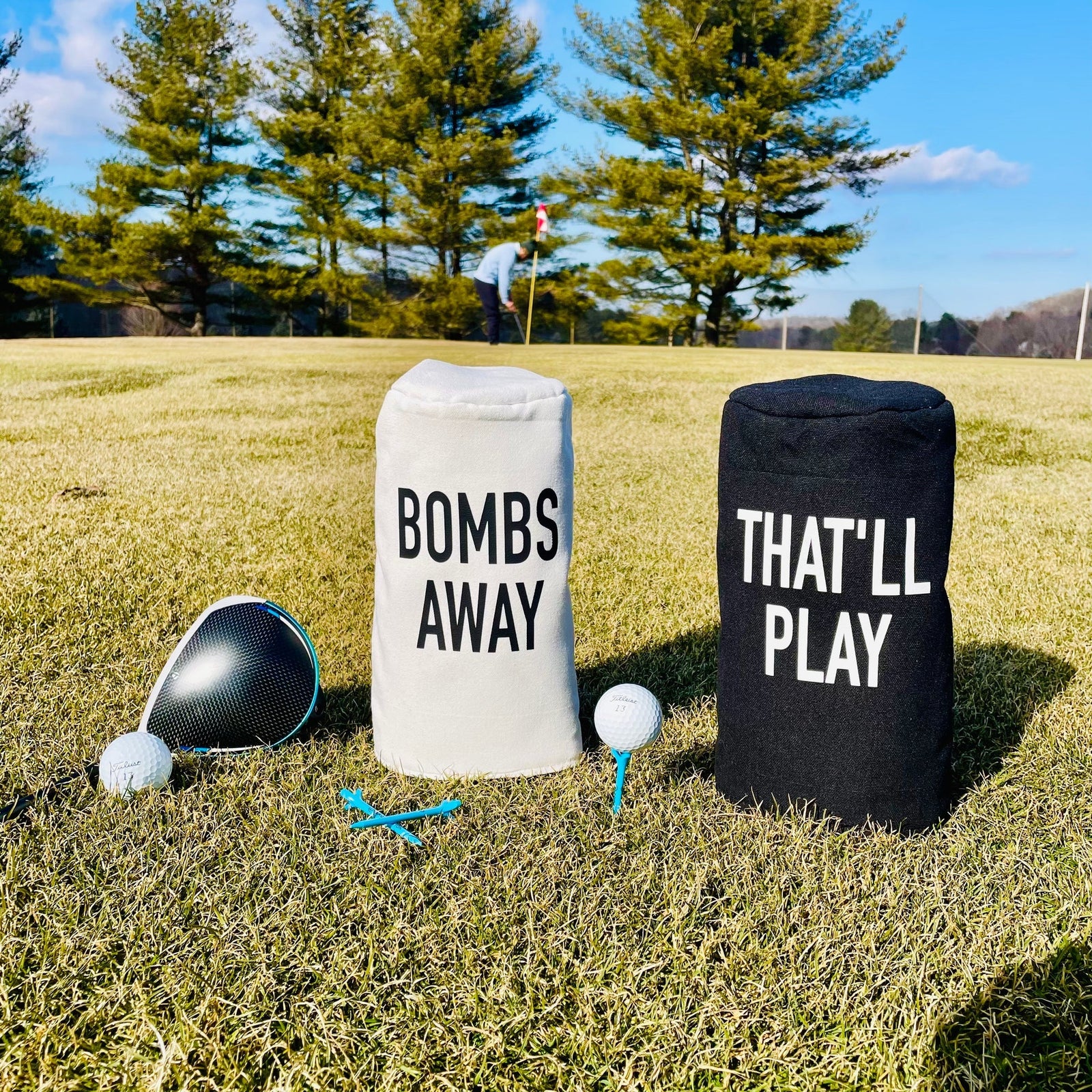Two golf barrel head covers on grass course, white cover printed "BOMBS AWAY" and black cover printed "THAT'LL PLAY," displayed with driver, balls, and tees