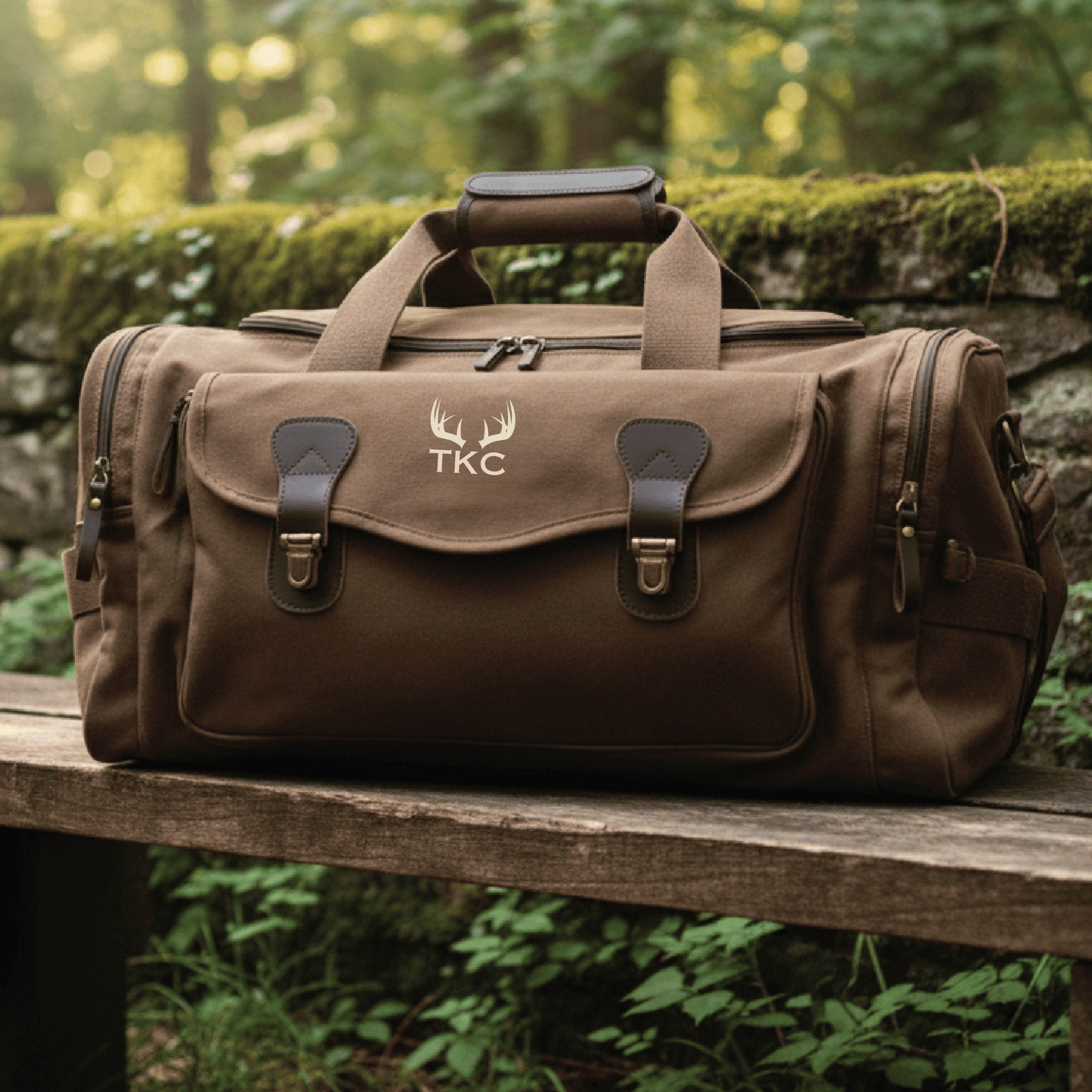 Brown canvas duffle bag with leather accents, embroidered with sample antler motif and "TKC" monogram on front flap, dual carry handles and side pockets visible