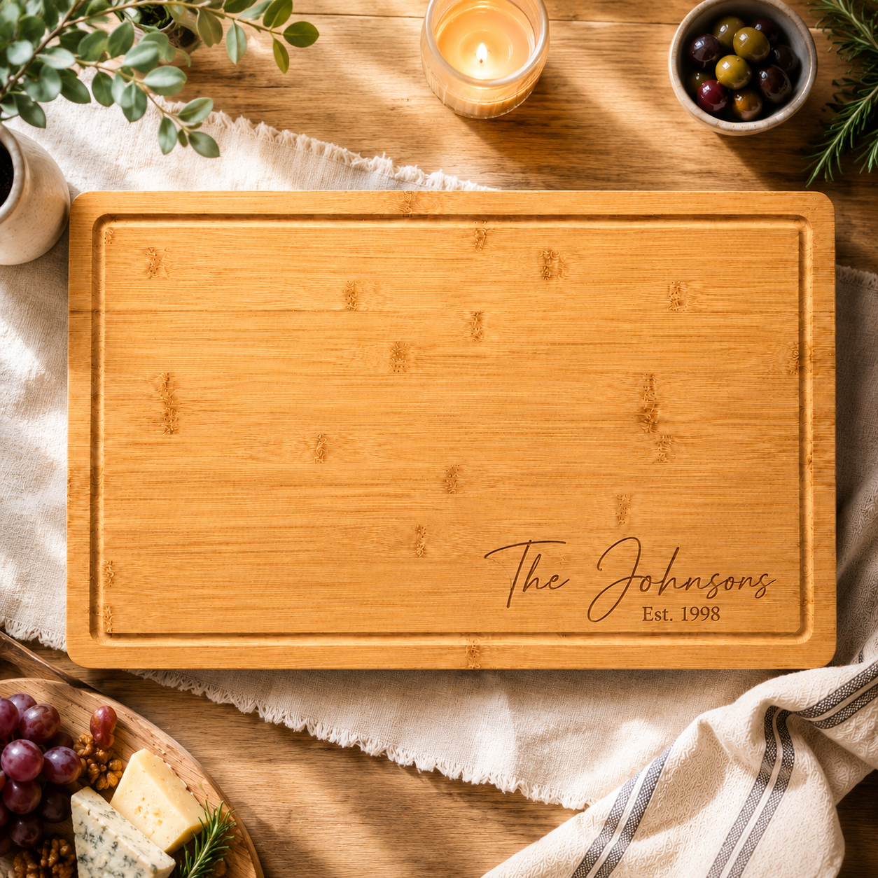 A rectangular bamboo serving board with rounded corners and engraved text reading 'The Johnsons Est. 1998' at the bottom right, styled on a wooden table with a candle, olives, grapes, cheese, and a striped cloth.