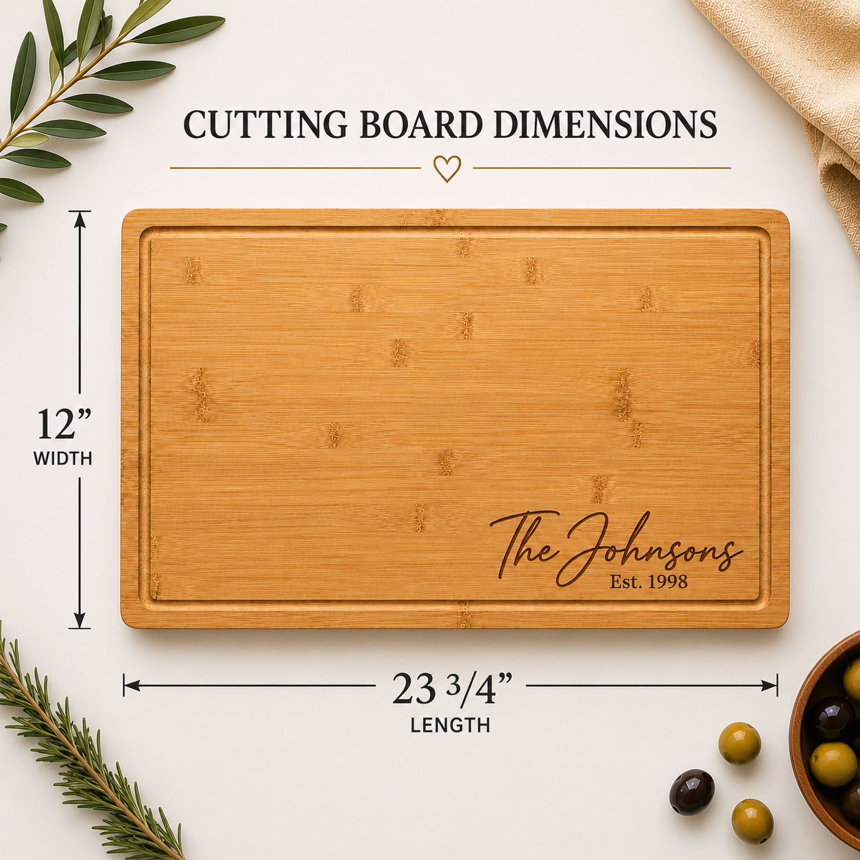 A rectangular bamboo cutting board with rounded corners and engraved script reading 'The Johnsons Est. 1998,' shown on a light neutral background with dimension callouts indicating 12 inches wide by 23 3/4 inches long.