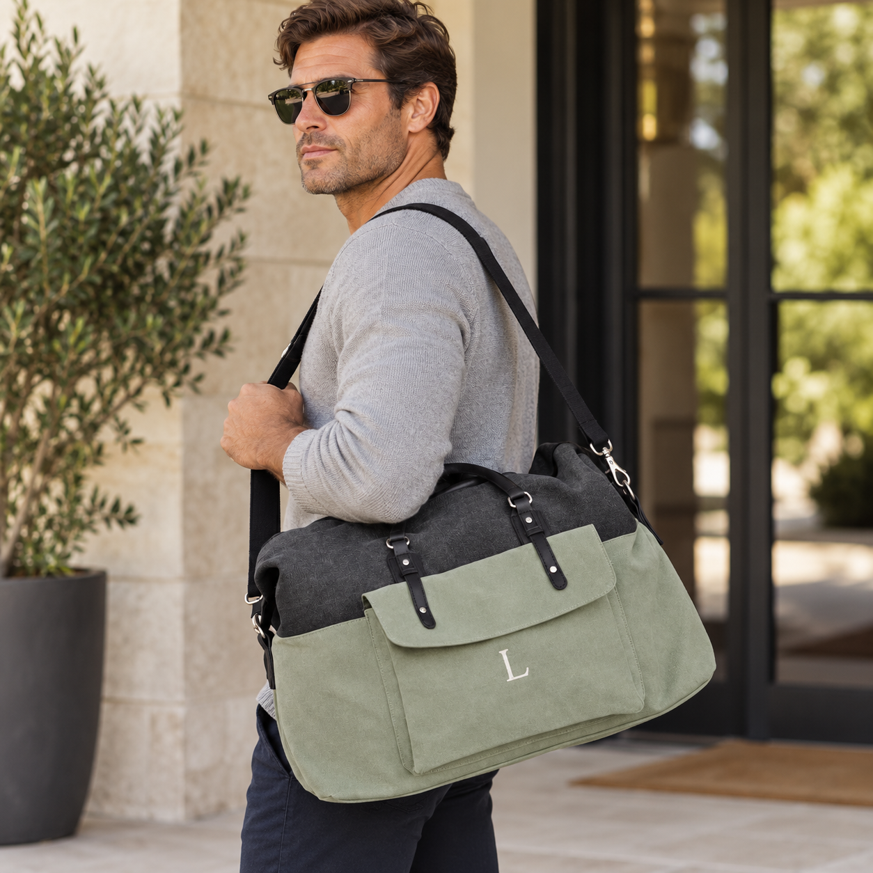 A man wearing sunglasses and a gray sweater carries a large two-tone duffel bag with an olive green front and dark gray top panel, featuring a silver monogram 'L' on the front pocket, in an outdoor patio setting.
