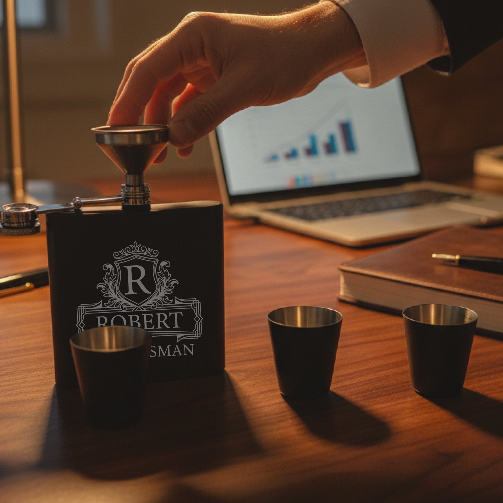 Black flask engraved with sample monogram "R ROBERT SMAN" and ornate crest, displayed on wooden desk with two matching shot glasses and funnel nearby