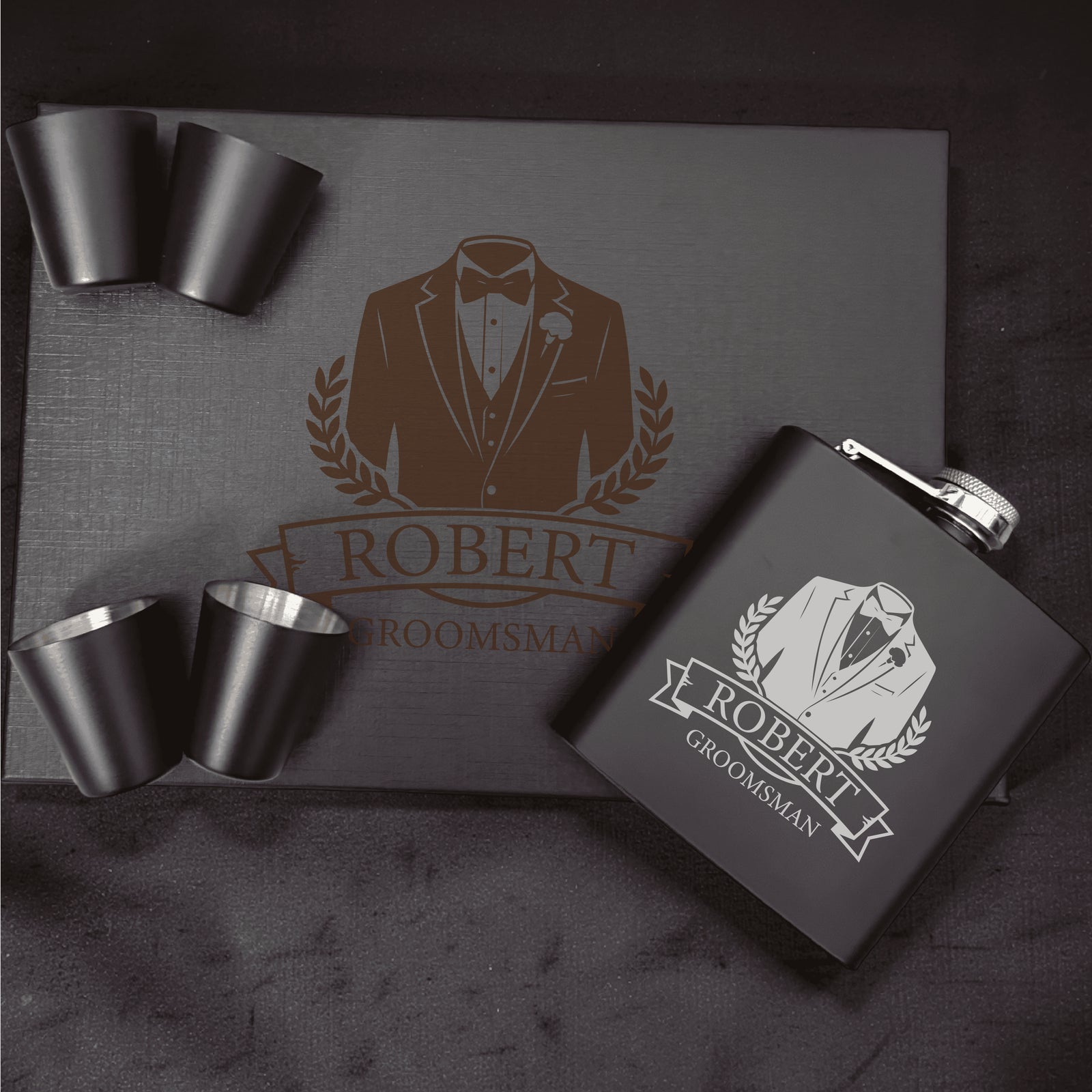 Flat lay of personalized flask gift set with black flask engraved "Robert Groomsman," gray box with matching tuxedo design, stainless steel shot glasses, and funnel on dark background