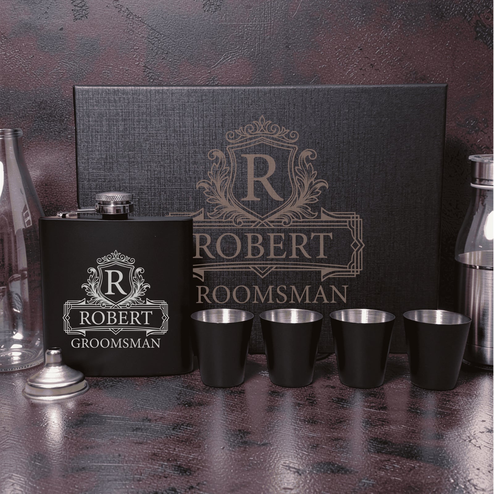 Black flask and four shot glasses with stainless steel rims, personalized with sample name "Robert" and "Groomsman" text in ornate crest design, displayed with matching gift box