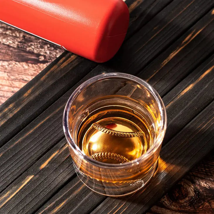 Whiskey glass with baseball-textured base containing amber liquid, positioned on dark wood surface next to red flask.