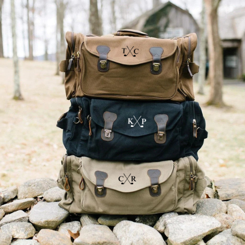 Three personalized duffels stacked showing sample monograms "IVC," "KYP," and "CXR" embroidered with crossed golf clubs motif in tan, navy, and cream canvas.