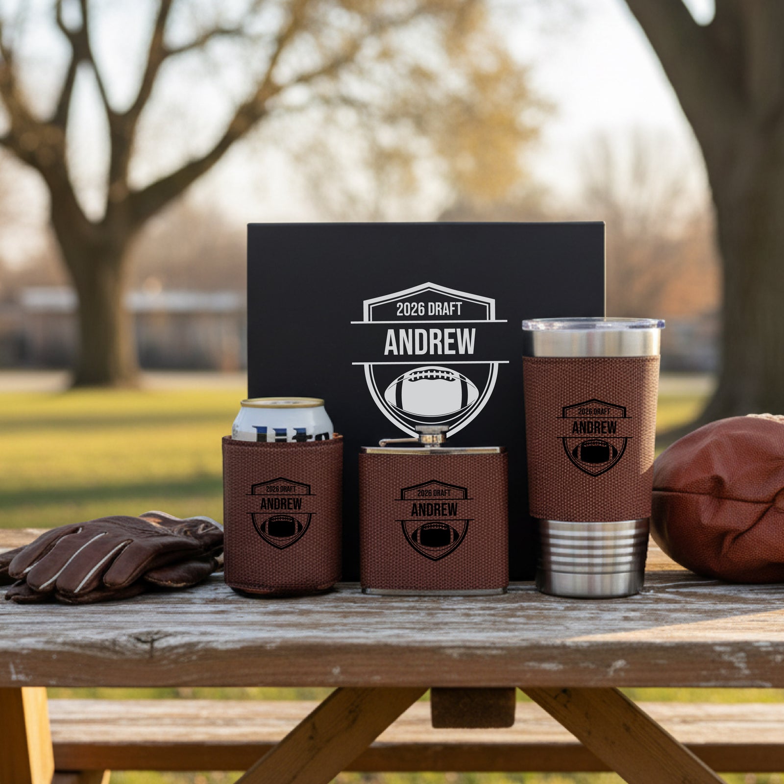 Brown leather koozies, can holder, and tumbler with brown gift box, all personalized with sample engraving "2026 DRAFT ANDREW" and football shield design, displayed outdoors on wooden table with football and gloves