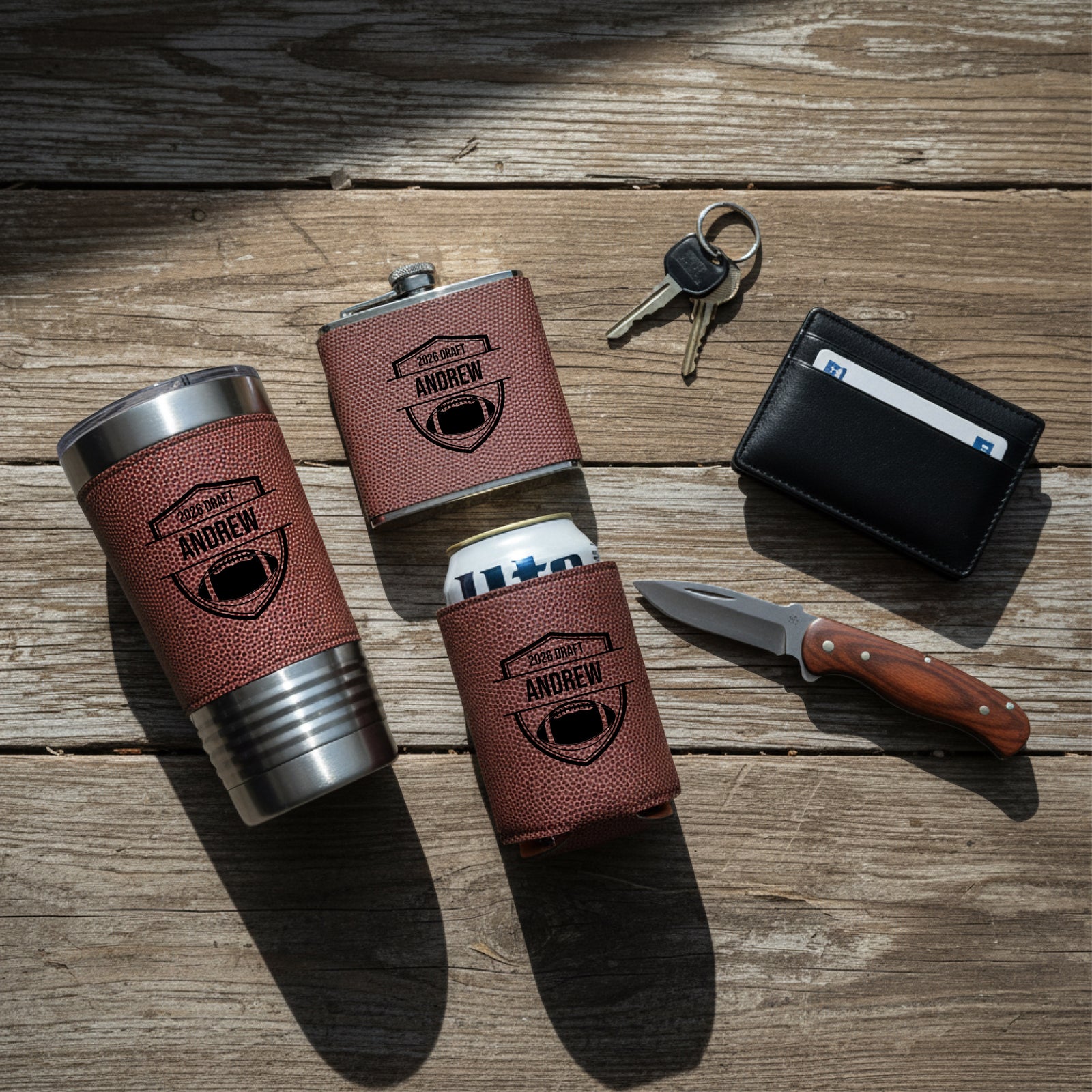 Lifestyle flatlay of personalized leather accessories with sample engraving "ANDREW" and football shield design, including insulated tumbler, flask, koozie, wallet, knife, and keys on wooden surface